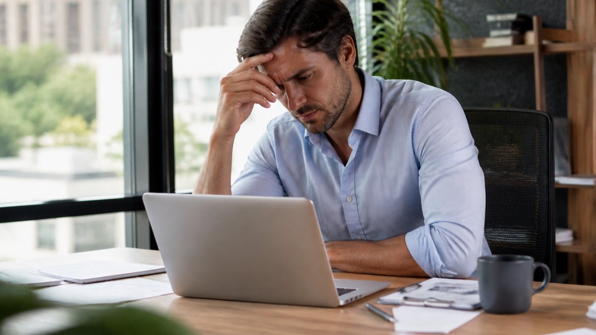 Person seated at a desk with a laptop, resting their forehead on one hand.